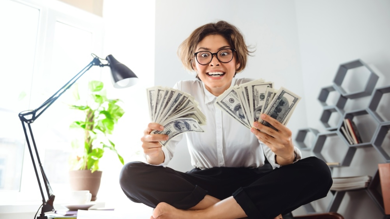 Young beautiful businesswoman in glasses smiling, holding money, sitting on table at workplace.