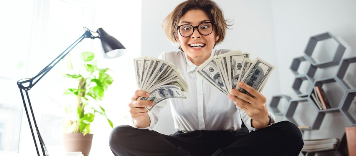 Young beautiful businesswoman in glasses smiling, holding money, sitting on table at workplace.